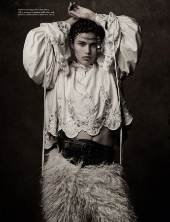 Model with braided hair wearing a white embroidered blouse and furry skirt with hands raised behind head