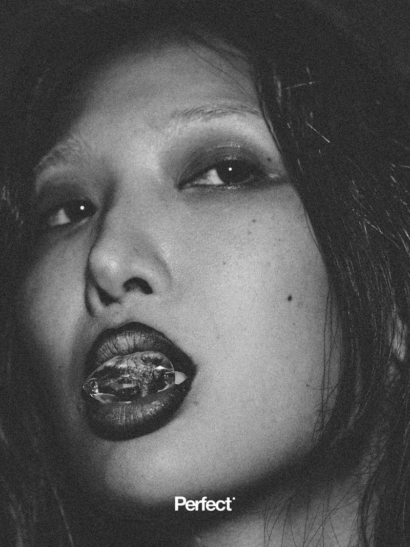 Close-up monochrome portrait of a person with dark hair holding a clear faceted stone between their lips
