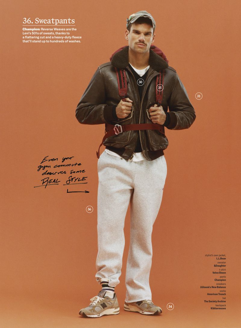 Man wearing a brown leather jacket, grey sweatpants, and cap standing against a warm solid background for GQ Magazine