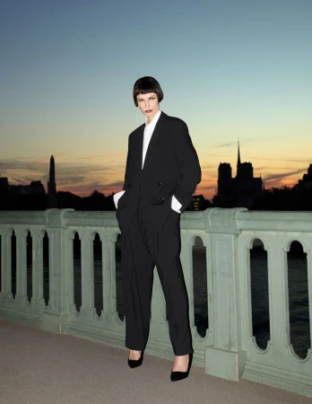 Natasha Poly wearing an oversized black suit standing on a bridge at sunset