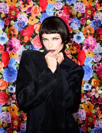 Natasha Poly wearing a dark structured coat against a vibrant floral backdrop for Pop Magazine
