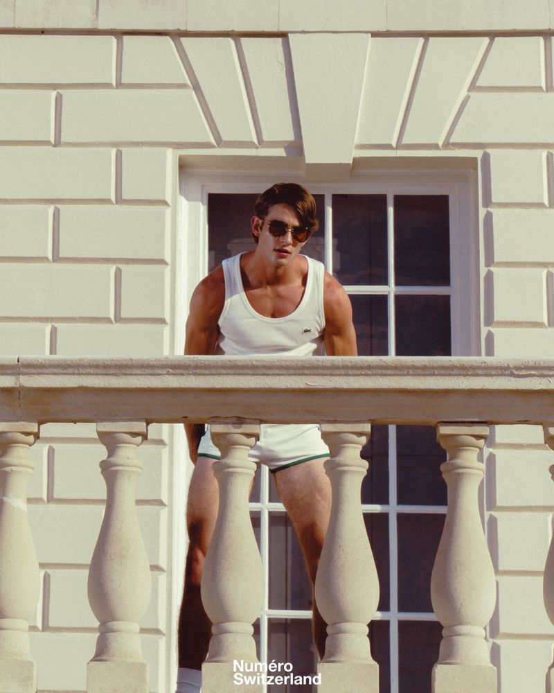 Man wearing sunglasses and a white tank top leaning over a stone balcony railing against a classical building exterior.