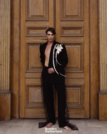 Male model wearing a black jacket and trousers standing before ornate wooden double doors for Numéro Switzerland.
