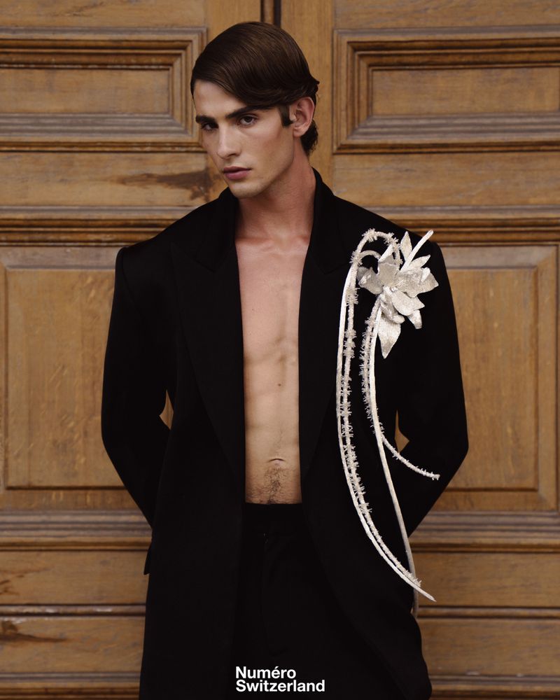 Portrait of a male model wearing an open black blazer with a white floral lapel accessory against a wooden door background.