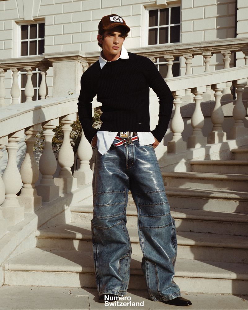 Young man in a black sweater and shiny blue jeans standing on classical stone stairs wearing a brown cap
