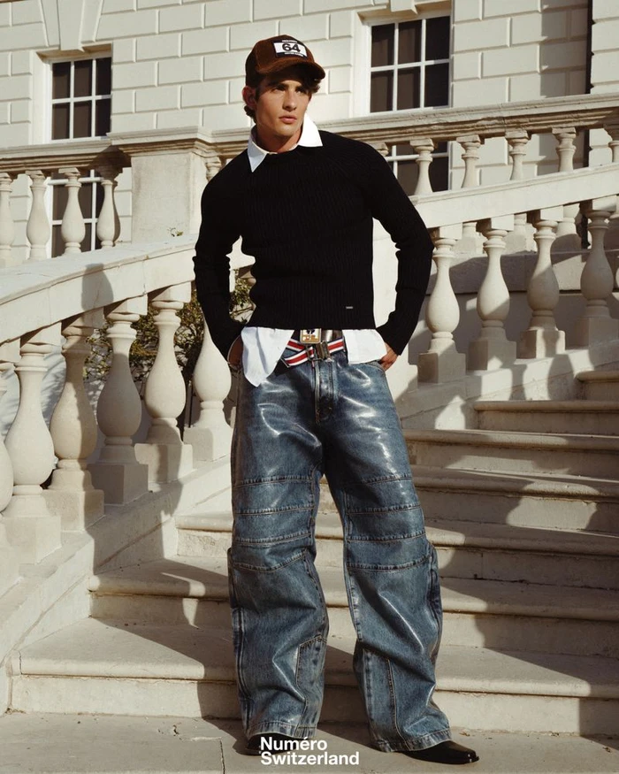 Young man in a black sweater and shiny blue jeans standing on classical stone stairs wearing a brown cap