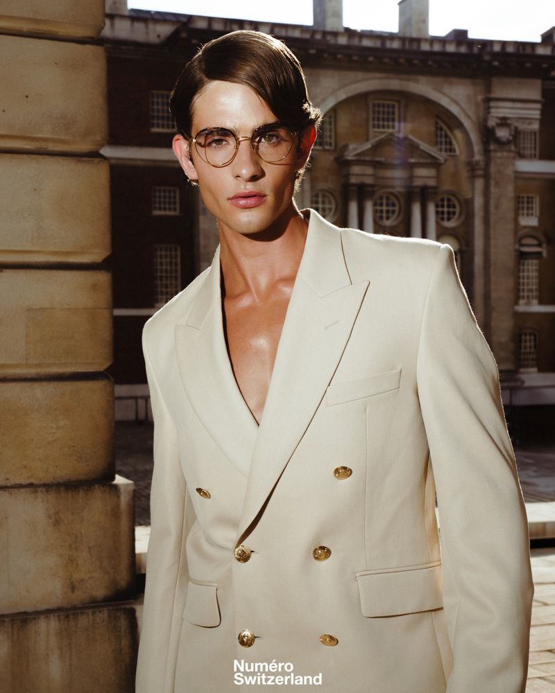 Man wearing a cream double-breasted blazer and round glasses outdoors in front of a classical building.