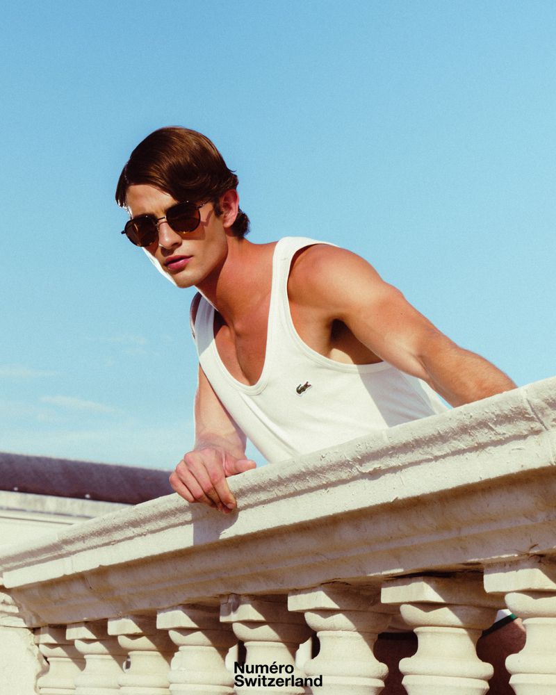 Man wearing sunglasses and a white tank top leaning over a decorative stone balcony against a clear blue sky