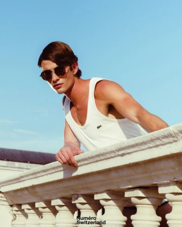 Man wearing sunglasses and a white tank top leaning over a decorative stone balcony against a clear blue sky