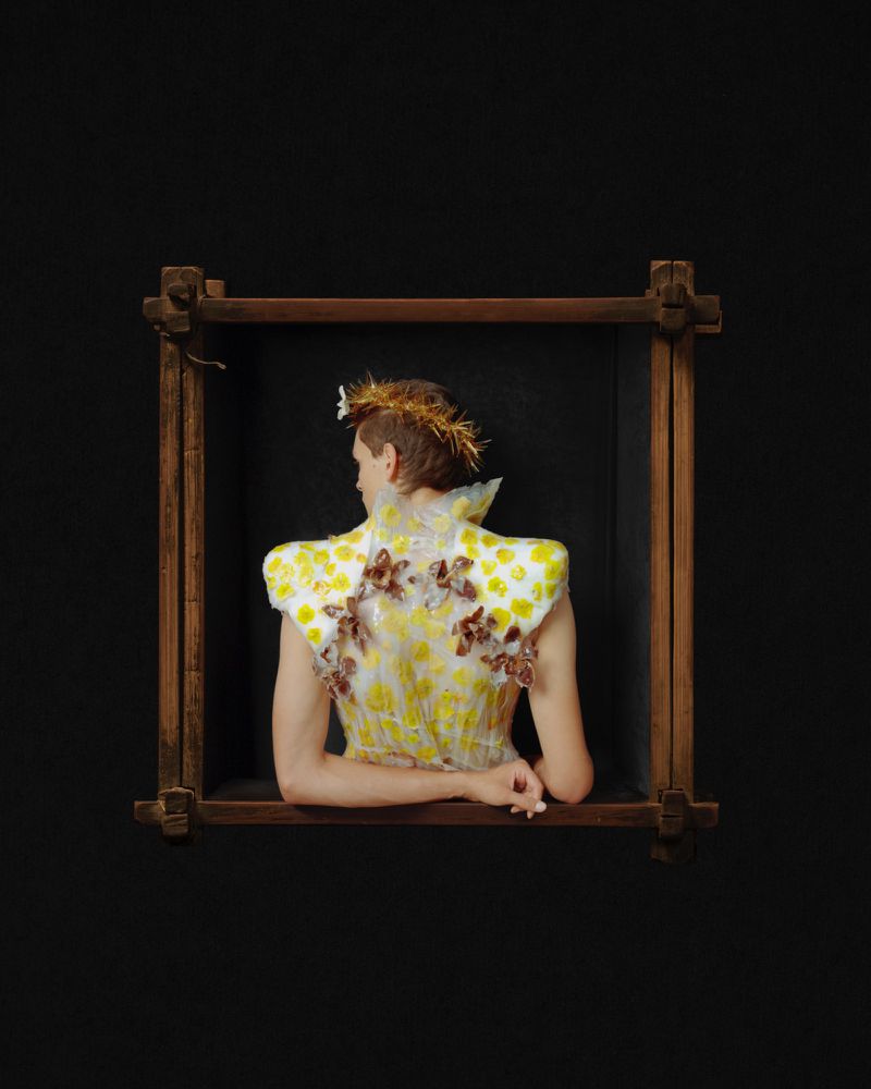 Model seen from behind wearing an elaborate floral-adorned garment, positioned inside a wooden frame against a black background.