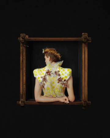Model seen from behind wearing an elaborate floral-adorned garment, positioned inside a wooden frame against a black background.