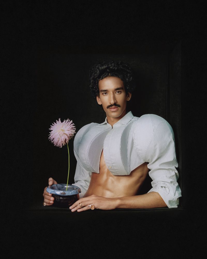 A man wearing a sculptural striped shirt holding a small vase with a pink flower against a black backdrop
