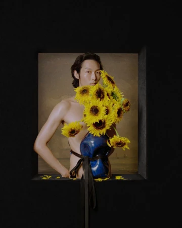 Shirtless male model holding a bouquet of yellow sunflowers covering his face, framed against a dark backdrop.