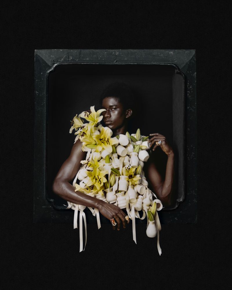 Model framed against a dark background wearing a sculptural garment of white vases and yellow lilies.