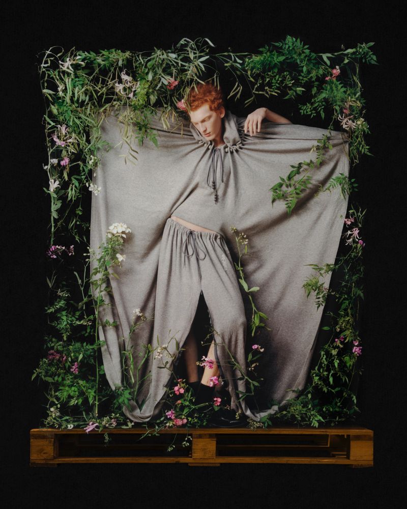 Model in grey hooded garment posing within a wooden frame draped with green foliage against a black background