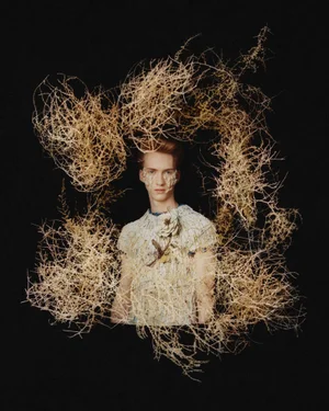Model surrounded by a wreath of dried branches wearing a textured white garment against a black background