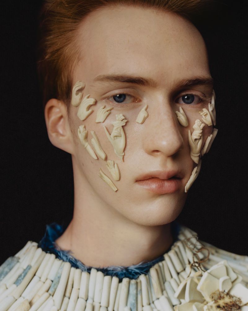 Close-up portrait of a young man with small ivory hand sculptures adhered to his face and wearing a beaded collar against a dark background.