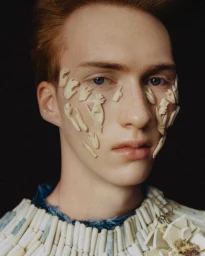 Close-up portrait of a young man with small ivory hand sculptures adhered to his face and wearing a beaded collar against a dark background.