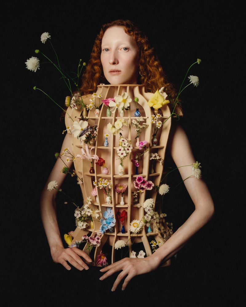 Model wearing a structured wooden grid corset filled with miniature vases and flowers against a dark background.