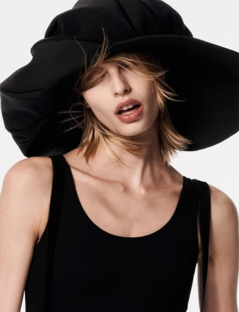 Model wearing a large black hat and black sleeveless top against a plain white background in an editorial portrait.