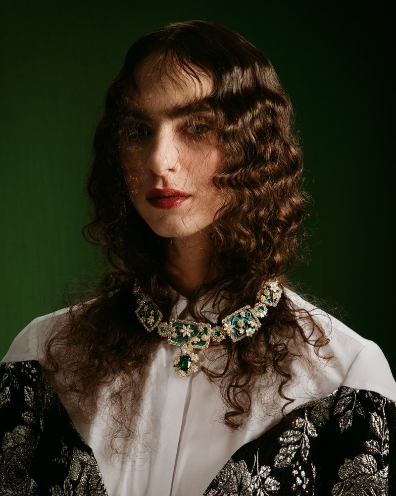 Portrait of a person with curly brown hair wearing a large green gem necklace, collared shirt, and patterned jacket against a green background.