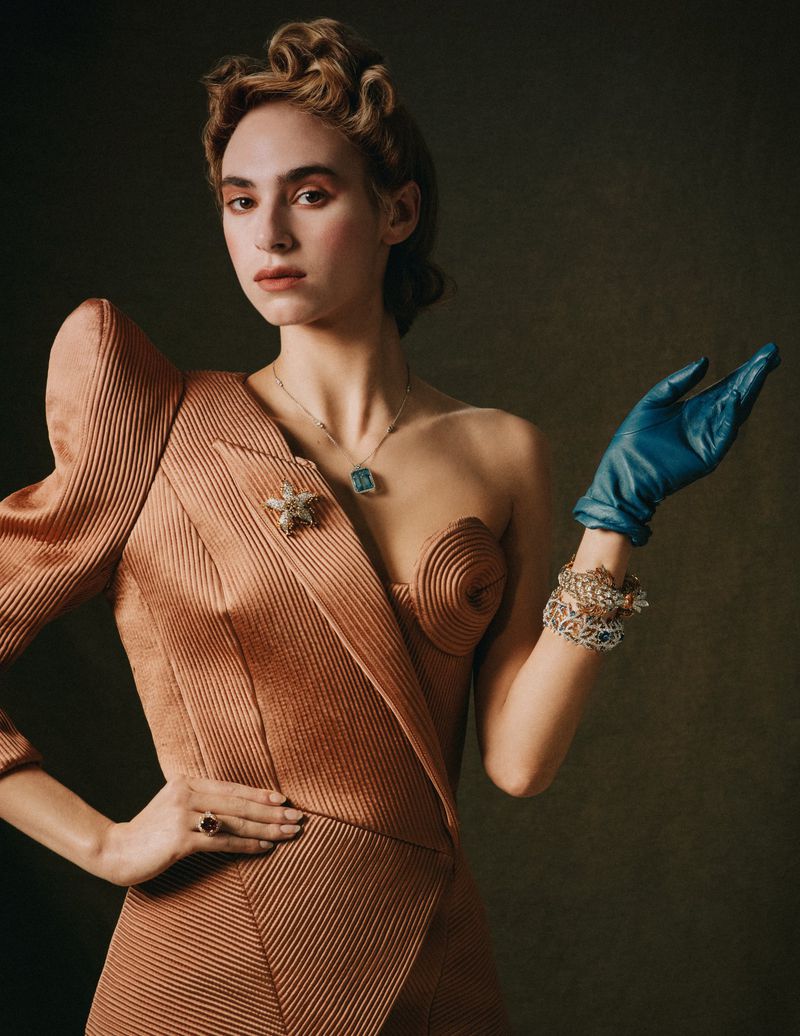 Elegant woman in peach dress with blue gloves posing in a studio, showcasing vintage fashion and jewelry against a muted backdrop.