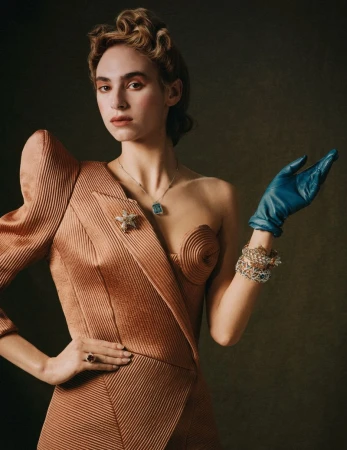 Elegant woman in peach dress with blue gloves posing in a studio, showcasing vintage fashion and jewelry against a muted backdrop.