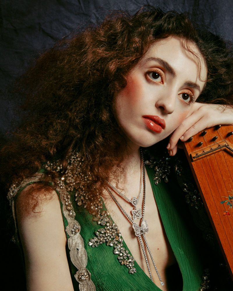 Portrait of a woman with curly auburn hair and pale skin, wearing a green beaded dress and silver jewelry while holding a stringed instrument.