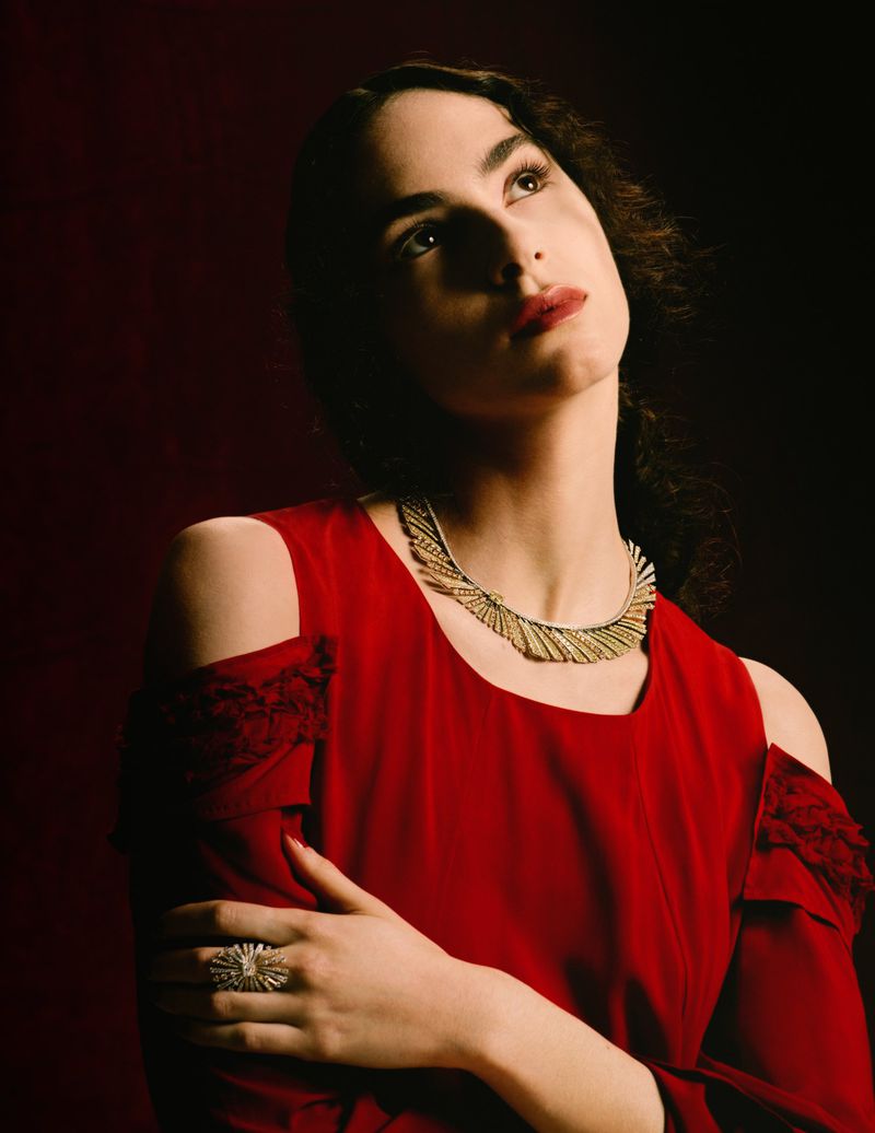 Portrait of a woman with dark hair wearing a red dress and ornate gold necklace and ring, looking upward in a studio setting.