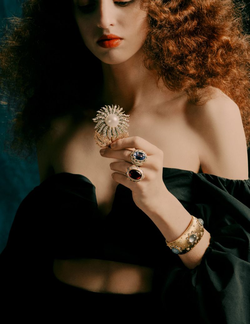 Elegant woman showcases vintage-inspired jewelry, including a golden cuff and gemstone rings, complemented by dramatic red lipstick and a dark dress.