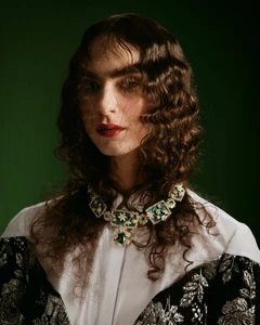 Model Clara Benador wearing an ornate green gemstone necklace and floral brocade jacket against a dark green studio backdrop.