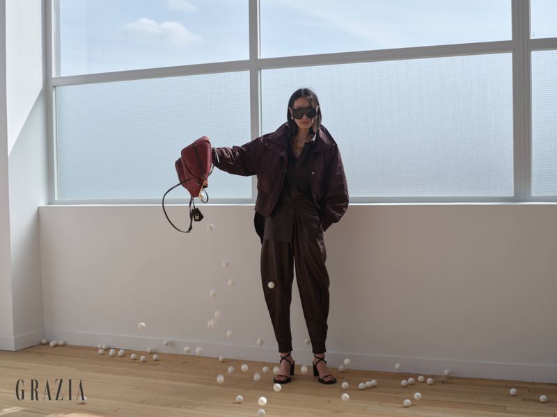 Model in brown leather jacket and trousers holding a red handbag spilling white spheres in front of large windows.