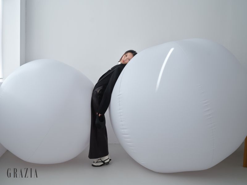 Model in black coat leaning between two large white spherical inflatables in a studio setting