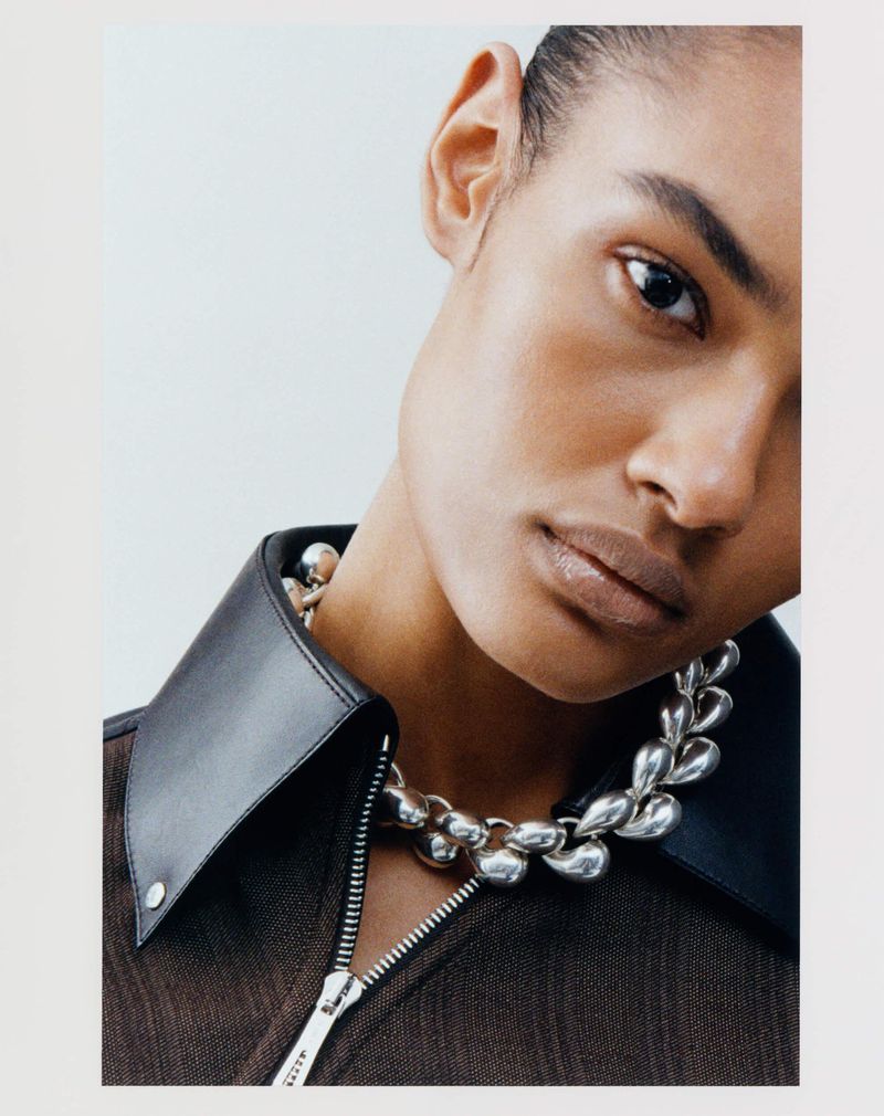 Close-up portrait of a model wearing a dark zipped collared top and silver statement necklace against a light background
