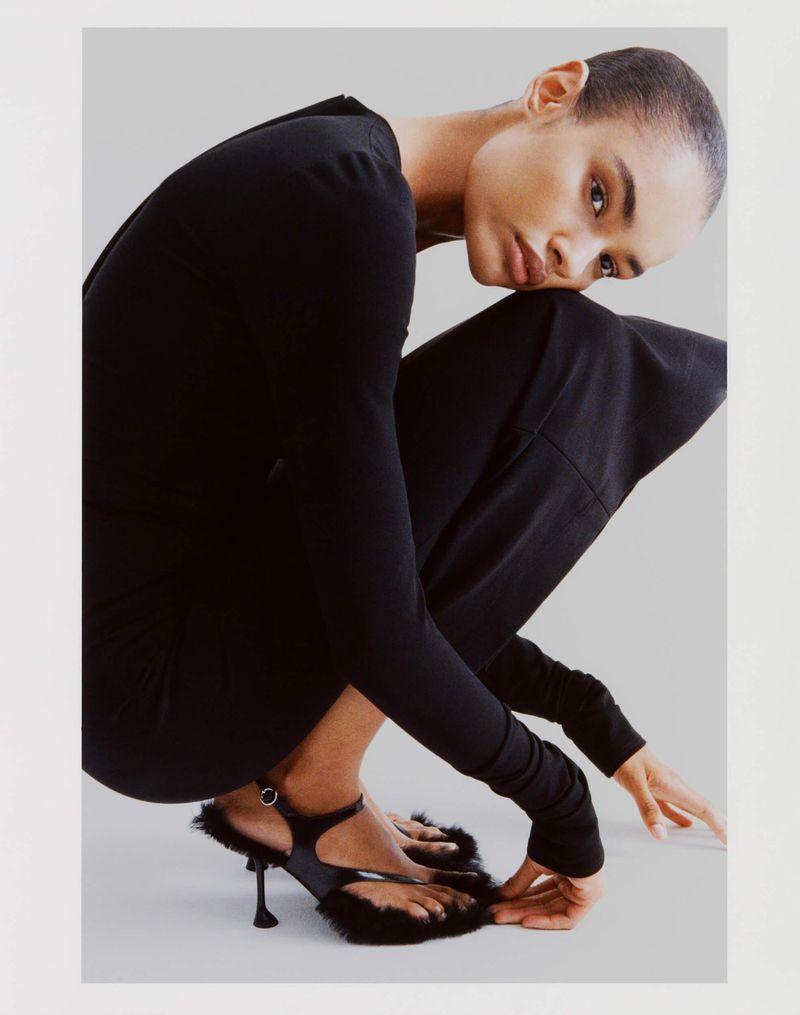 Model with a buzzcut wearing a black top and furry black heels, crouching against a neutral studio backdrop