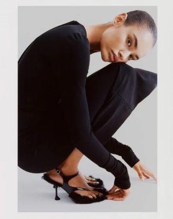 Model with a buzzcut wearing a black top and furry black heels, crouching against a neutral studio backdrop