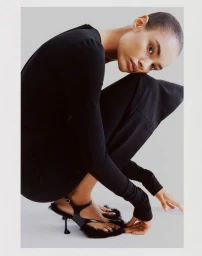 Model with a buzzcut wearing a black top and furry black heels, crouching against a neutral studio backdrop