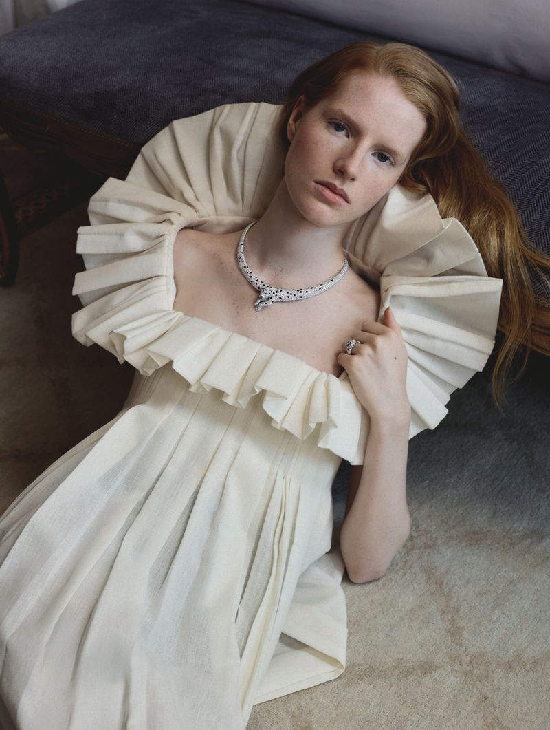 Model wearing a structured white ruffled dress and diamond panther necklace while reclining on a dark seat