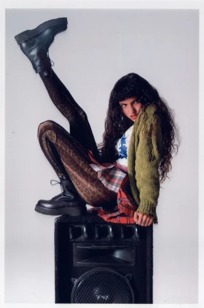A young woman with dark curly hair wearing a green cardigan, plaid skirt, and platform boots poses perched atop a black speaker cabinet.