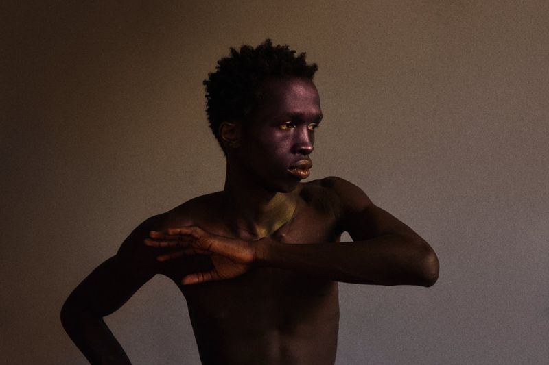 Portrait of a black male model posing with arms raised against a plain textured background.
