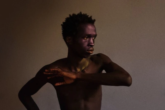 Portrait of a black male model posing with arms raised against a plain textured background.