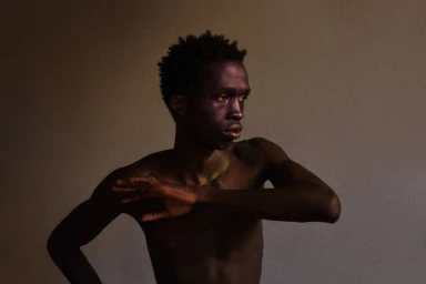 Portrait of a black male model posing with arms raised against a plain textured background.