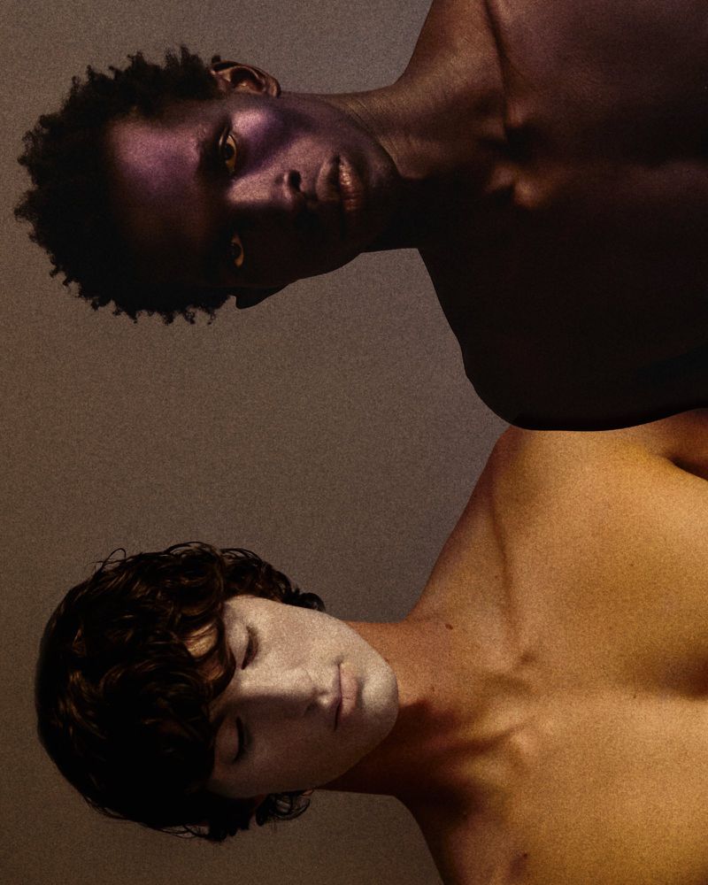Portrait of two male models with distinct skin and makeup tones against a textured neutral background