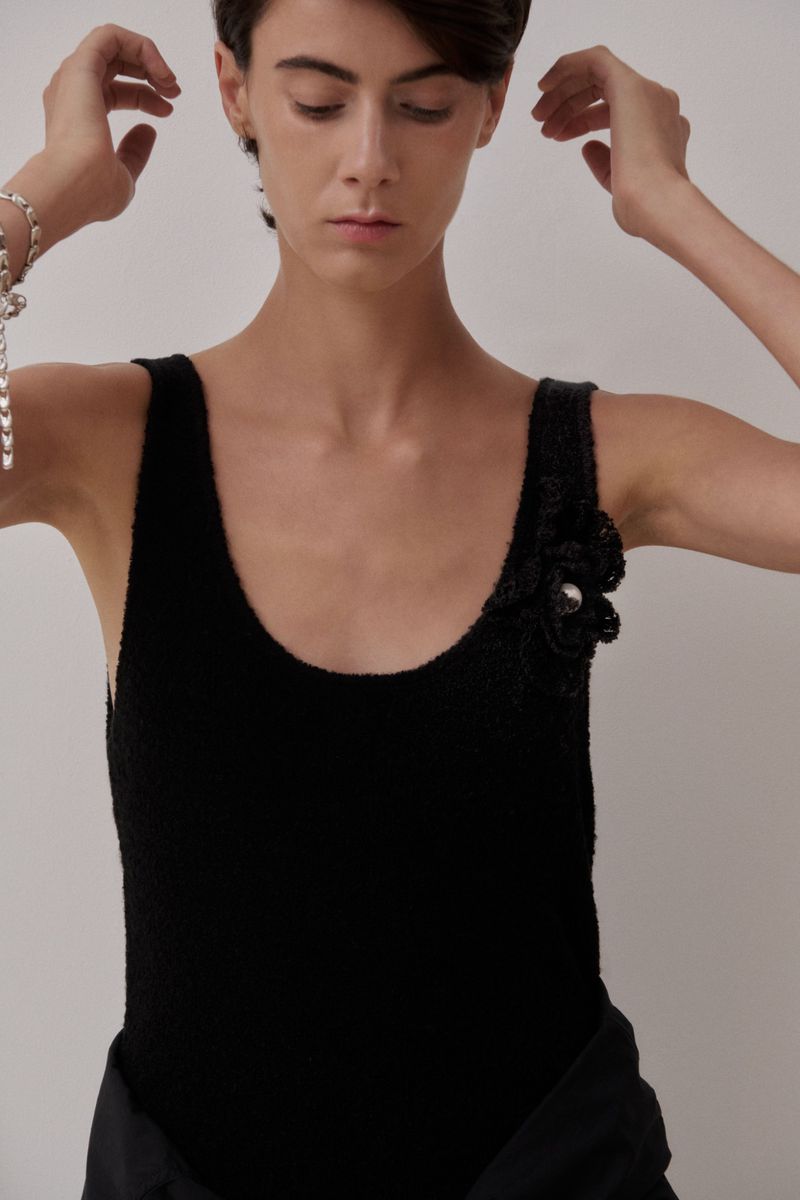 Model wearing a black scoop-neck tank top with a floral shoulder appliqué against a plain pale background