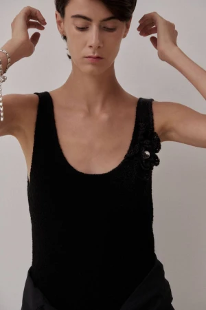 Model wearing a black scoop-neck tank top with a floral shoulder appliqué against a plain pale background