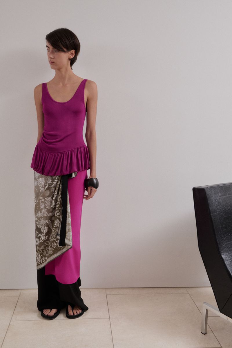 Model wearing magenta sleeveless top, patterned wrap skirt, and black trousers against a neutral interior background