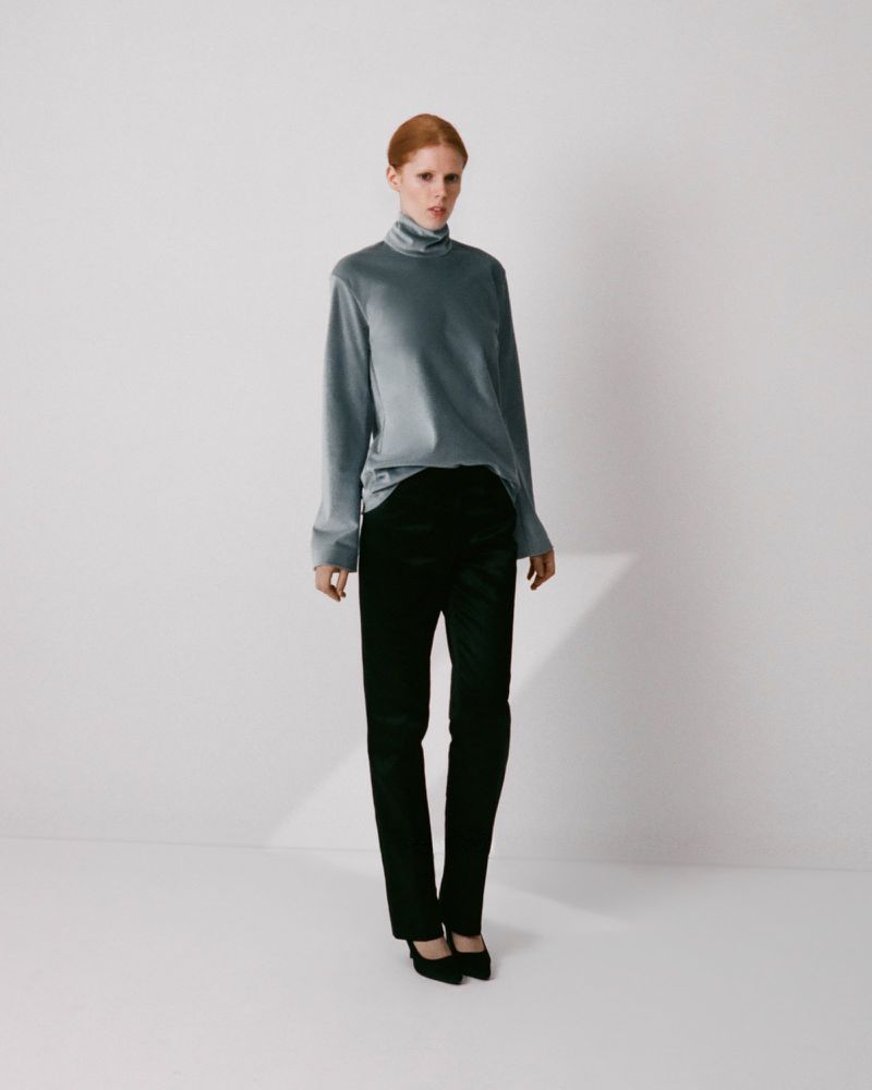 Model with red hair wearing a grey turtleneck top and black trousers against a neutral white background