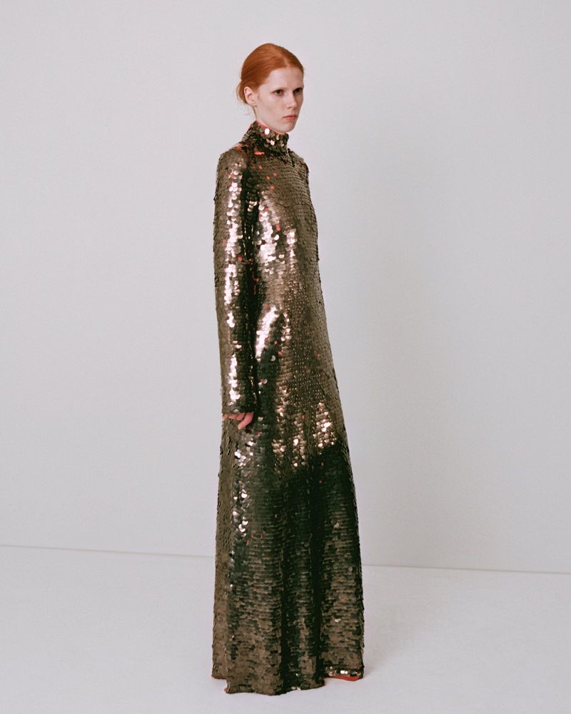 Model wearing a long metallic sequin dress standing against a plain white studio wall