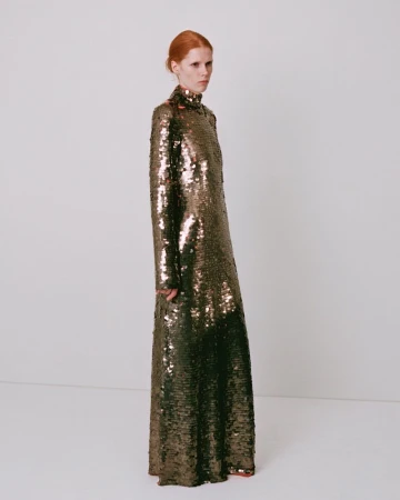 Model wearing a long metallic sequin dress standing against a plain white studio wall