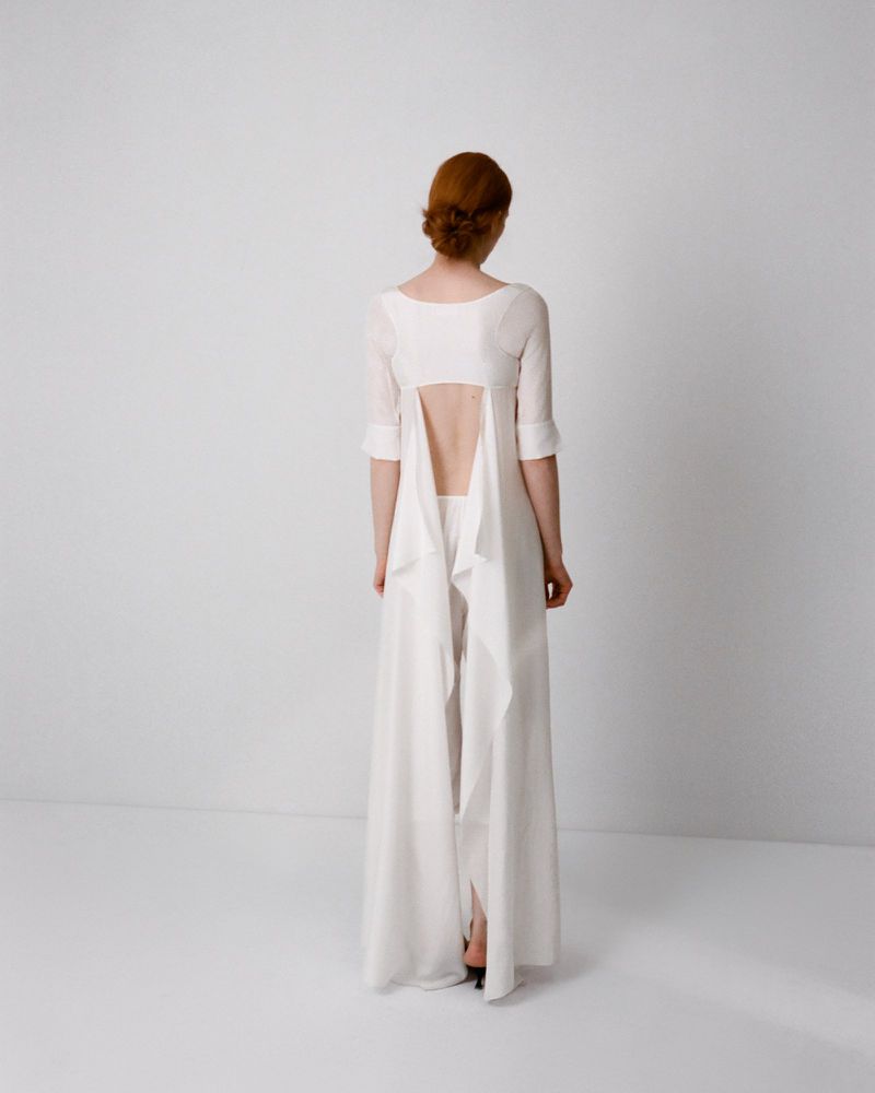 Model with red hair viewed from behind wearing a white open-back long dress against a plain white seamless studio background.